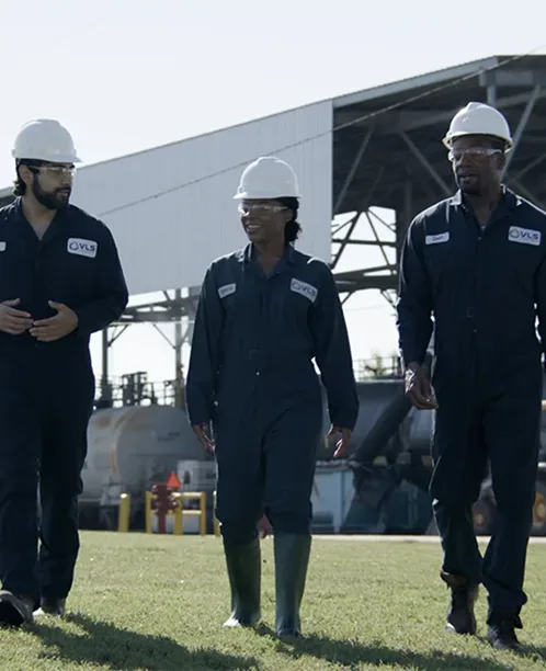 Three industrial workers in navy coveralls and white hard hats walking across a grassy area at a facility site.