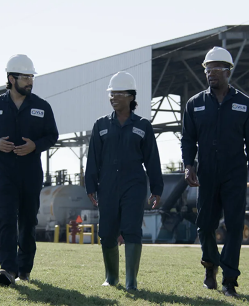 Three industrial workers in navy coveralls and white hard hats walking across a grassy area at a facility site.