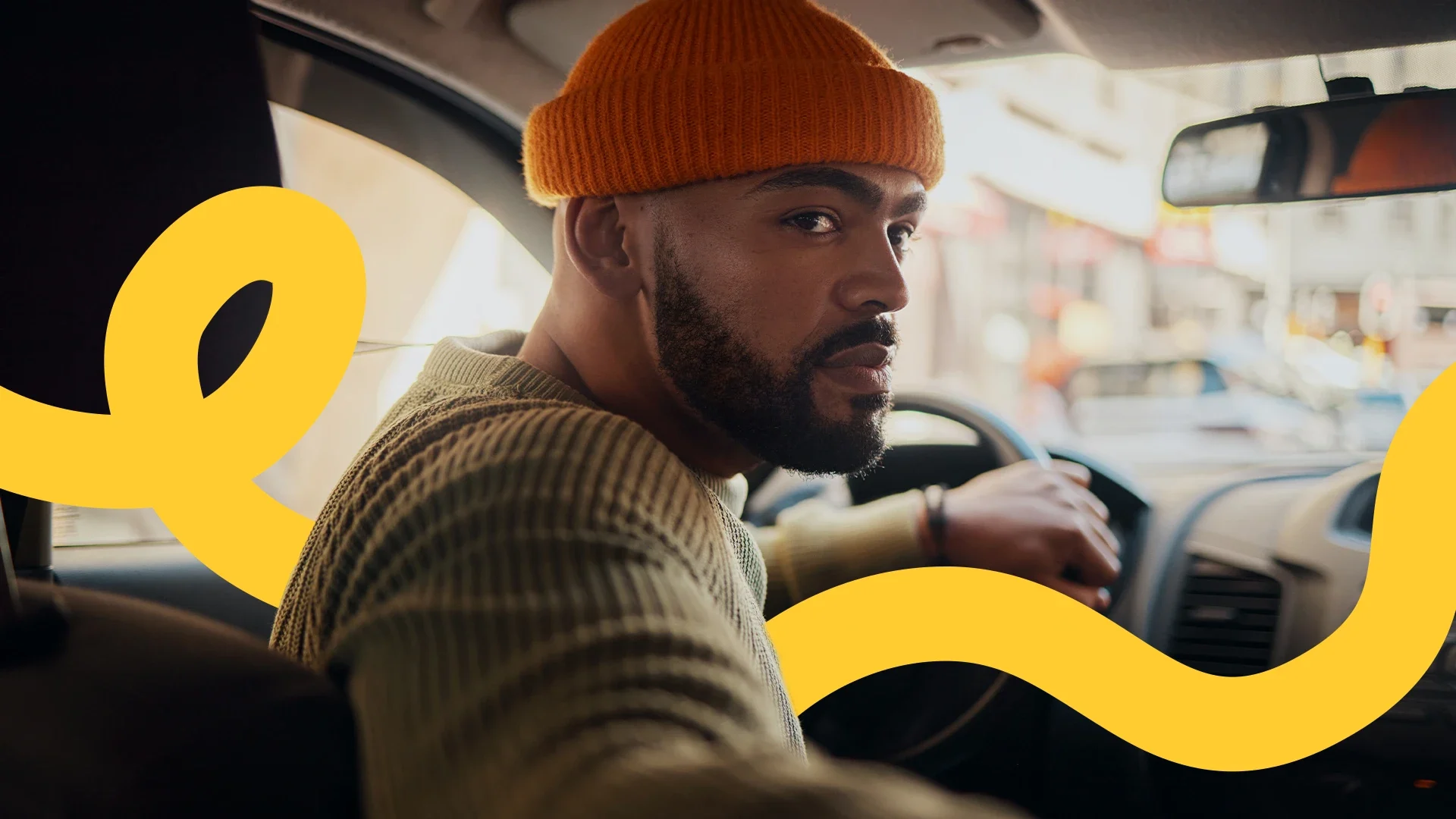 Mann mit orangefarbener Mütze und Strickpullover sitzt am Steuer eines Autos in der Stadt.