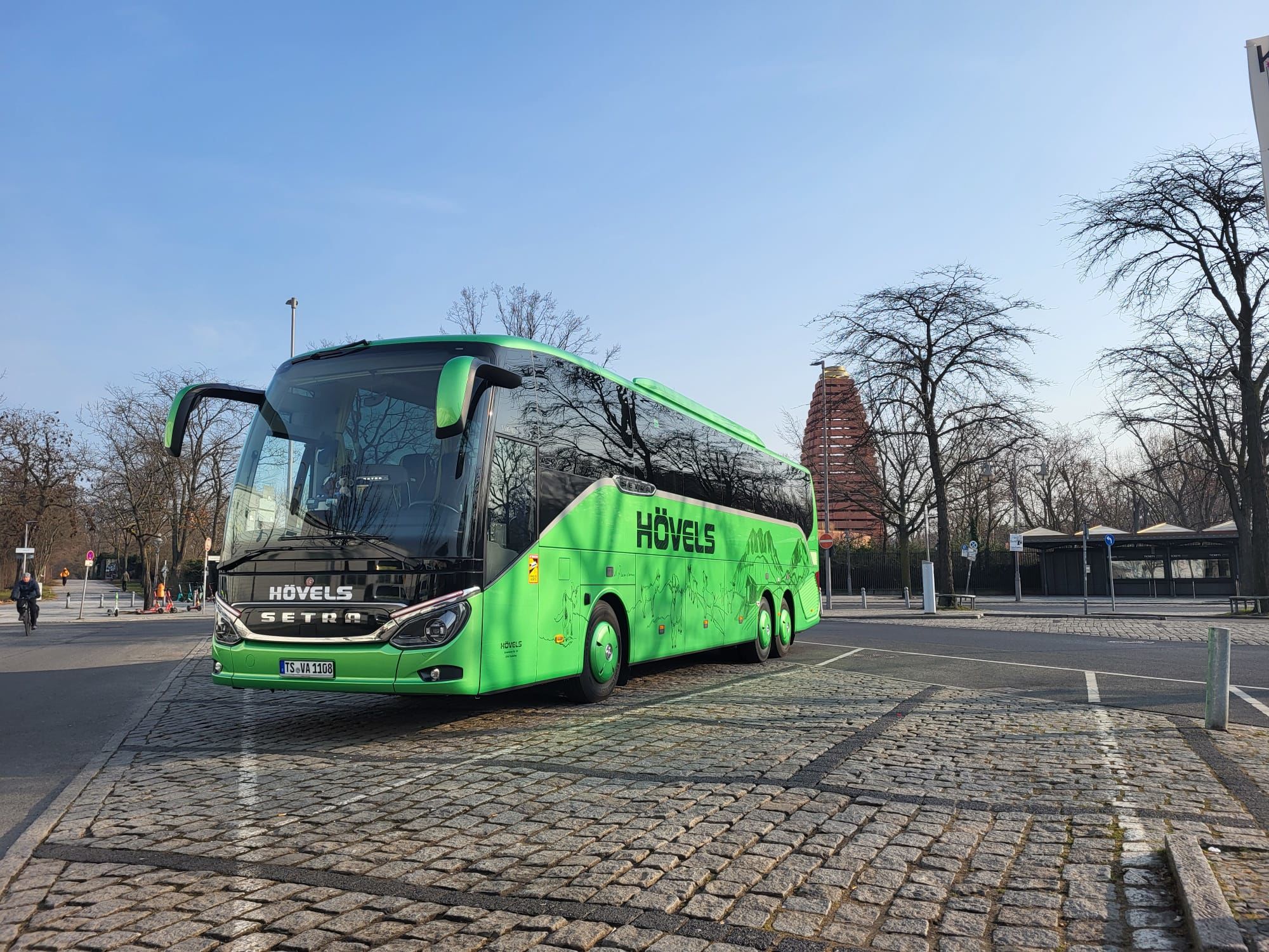 A green Setra coach bus branded "Hövels" parked outdoors at sunset, with its door open and a building in the background.
