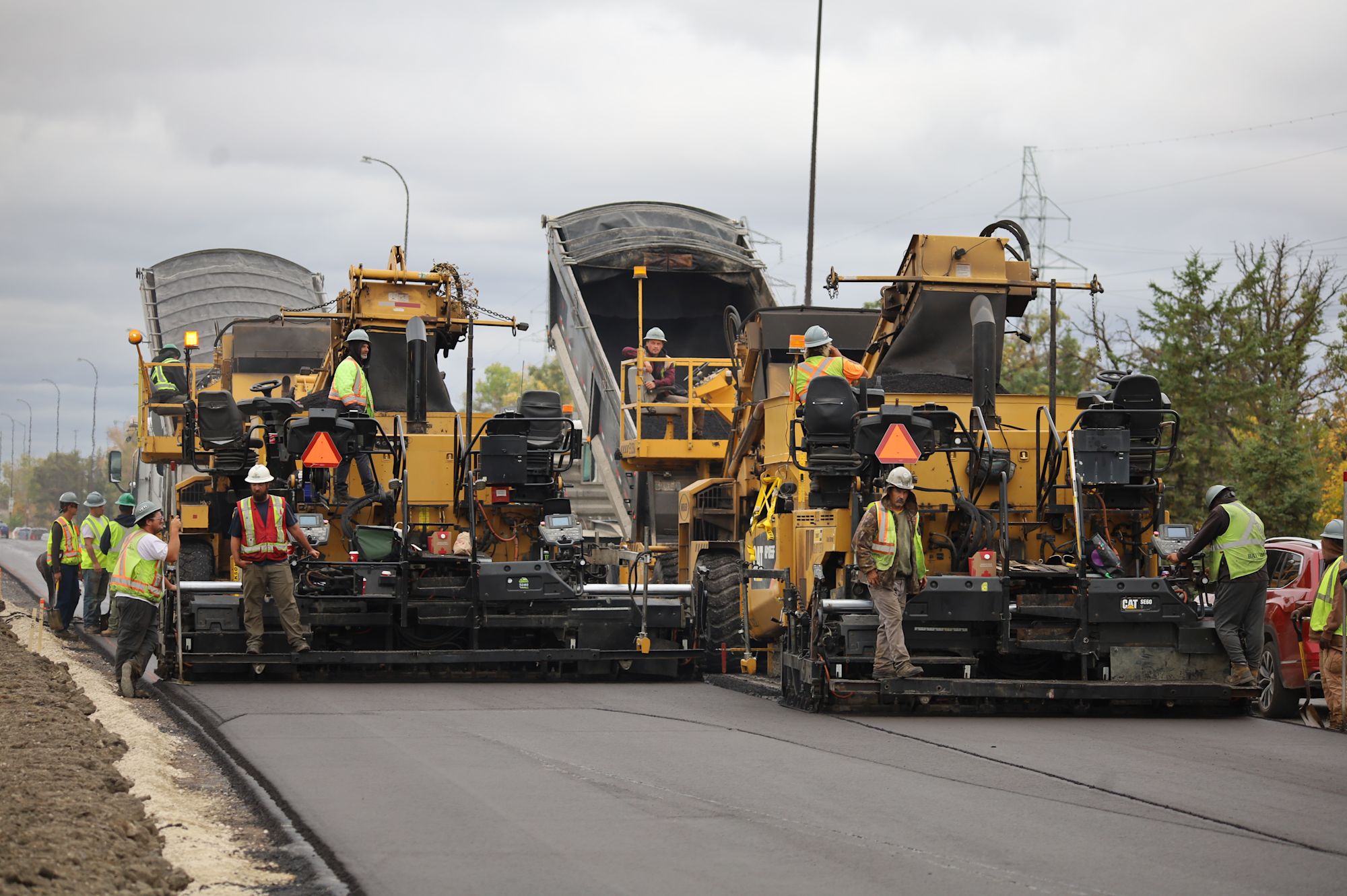 Construction workers in high-visibility vests operating yellow asphalt paving machines on a road under cloudy skies.