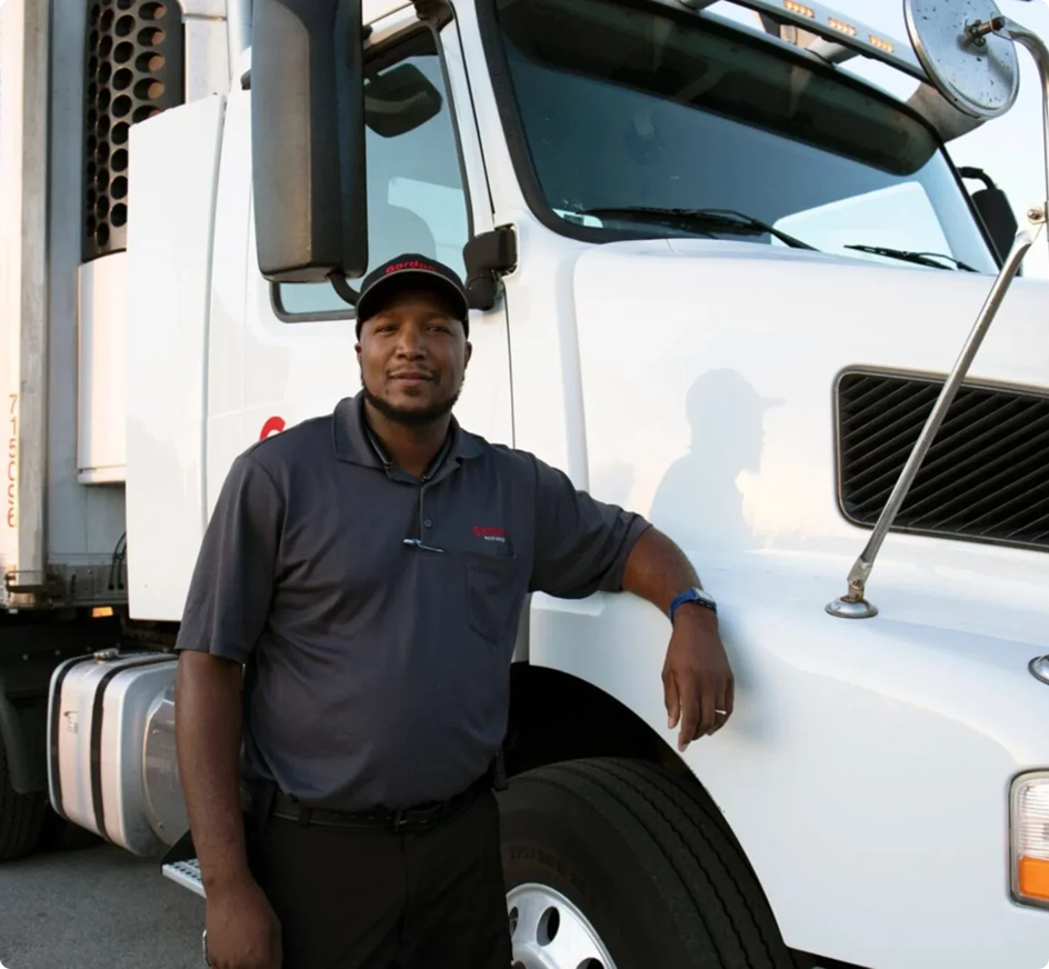 Professional truck driver in gray polo shirt and cap standing beside a white semi truck cab.