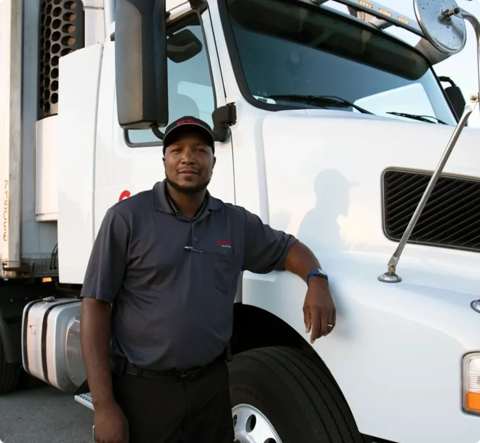 Professional truck driver in gray polo shirt and cap standing beside a white semi truck cab.