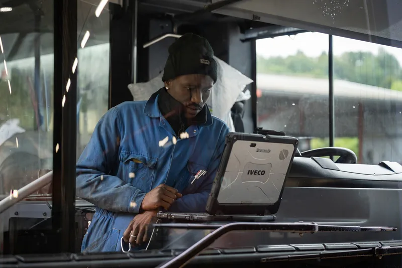 A technician in a blue jumpsuit and black beanie uses an Iveco rugged laptop inside a bus cabin.
