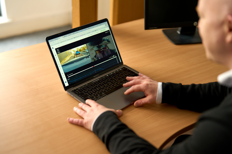 A person in a dark suit reviews dashcam footage on a laptop at a wooden desk, with a monitor visible in the background.