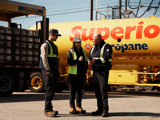 Three workers in safety vests discuss near a yellow Superior Propane tanker truck at an industrial site.
