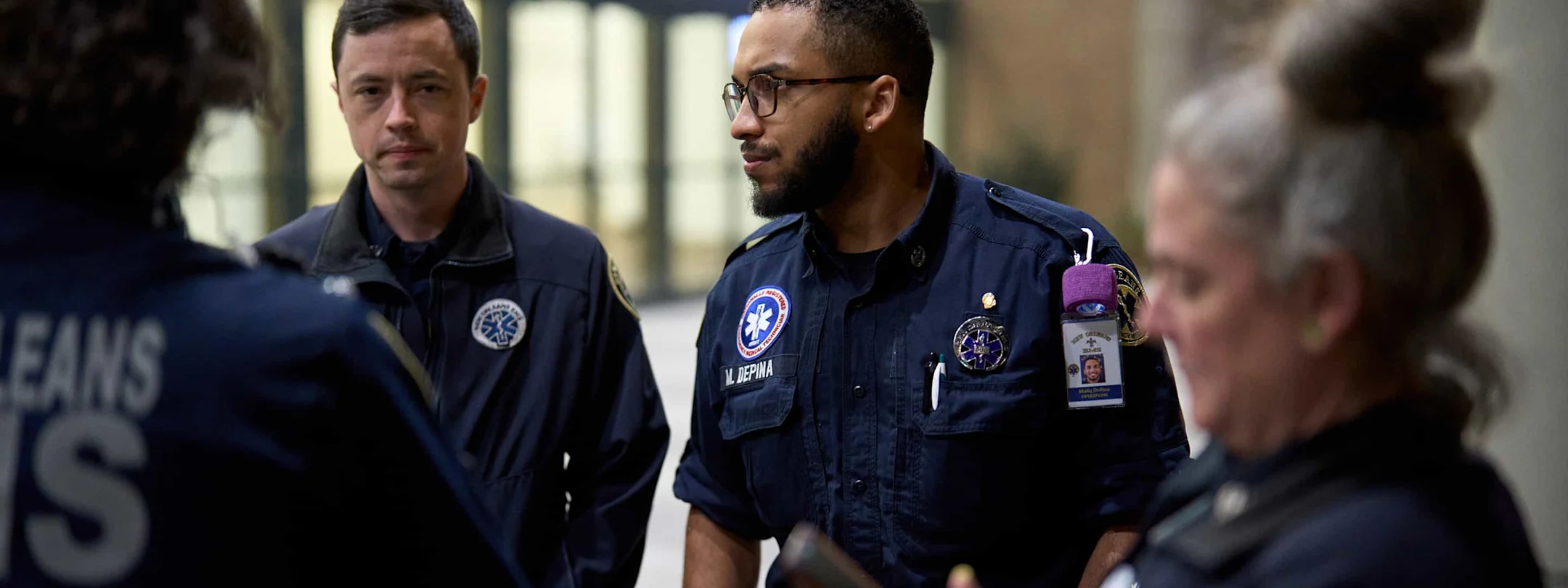 Two paramedics in navy uniforms with medical patches having a serious conversation in a hospital hallway.