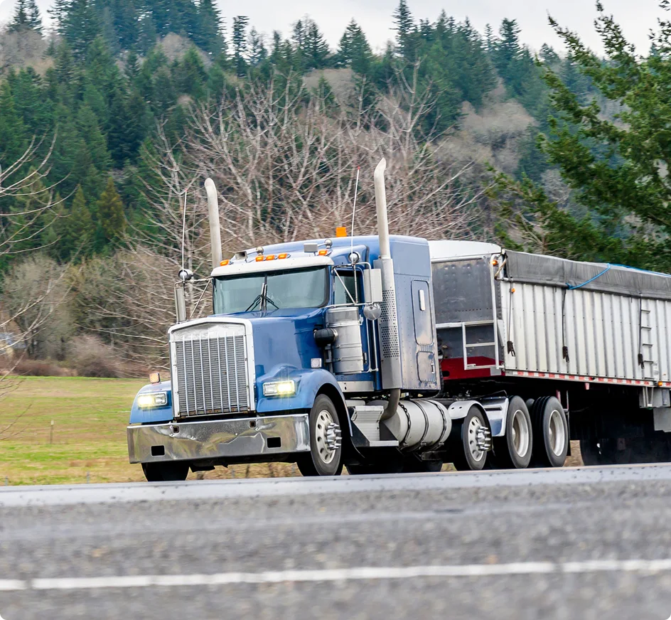 Blue semi truck with silver trailer driving on highway with evergreen forest backdrop