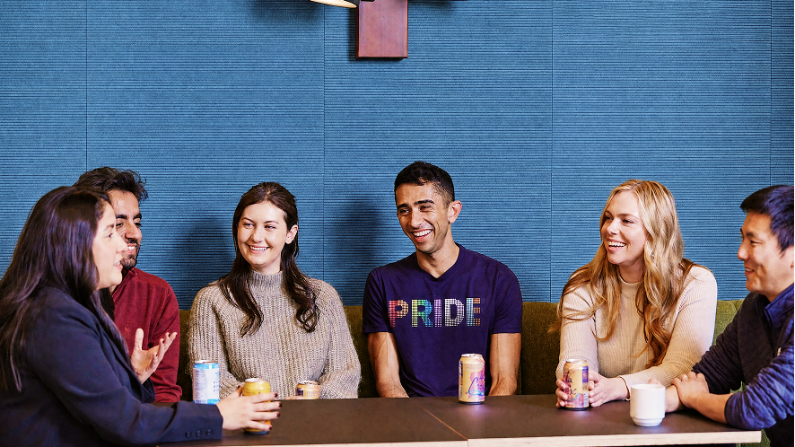 Six diverse colleagues laugh and chat around a table with drinks, one wearing a colorful Pride shirt, blue wall behind them.