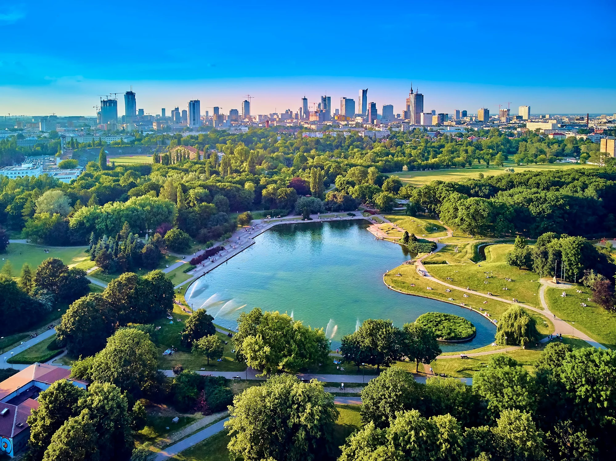Aerial view of a city park with a blue lake surrounded by green trees, with a modern skyline visible in the background.