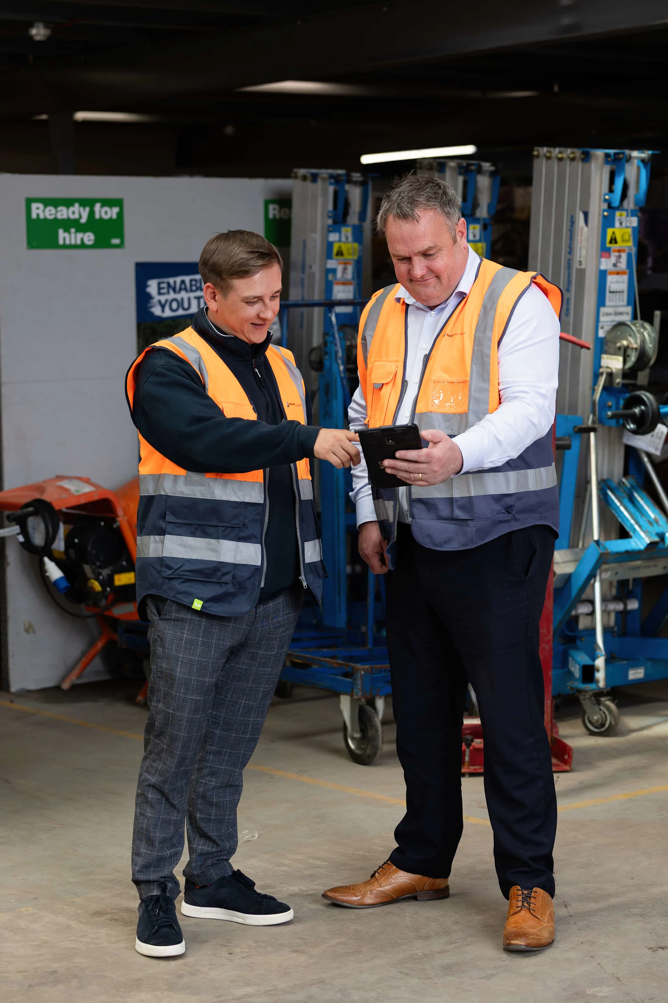 Deux ouvriers en gilets de sécurité orange consultent une tablette dans un entrepôt d'équipement de location.