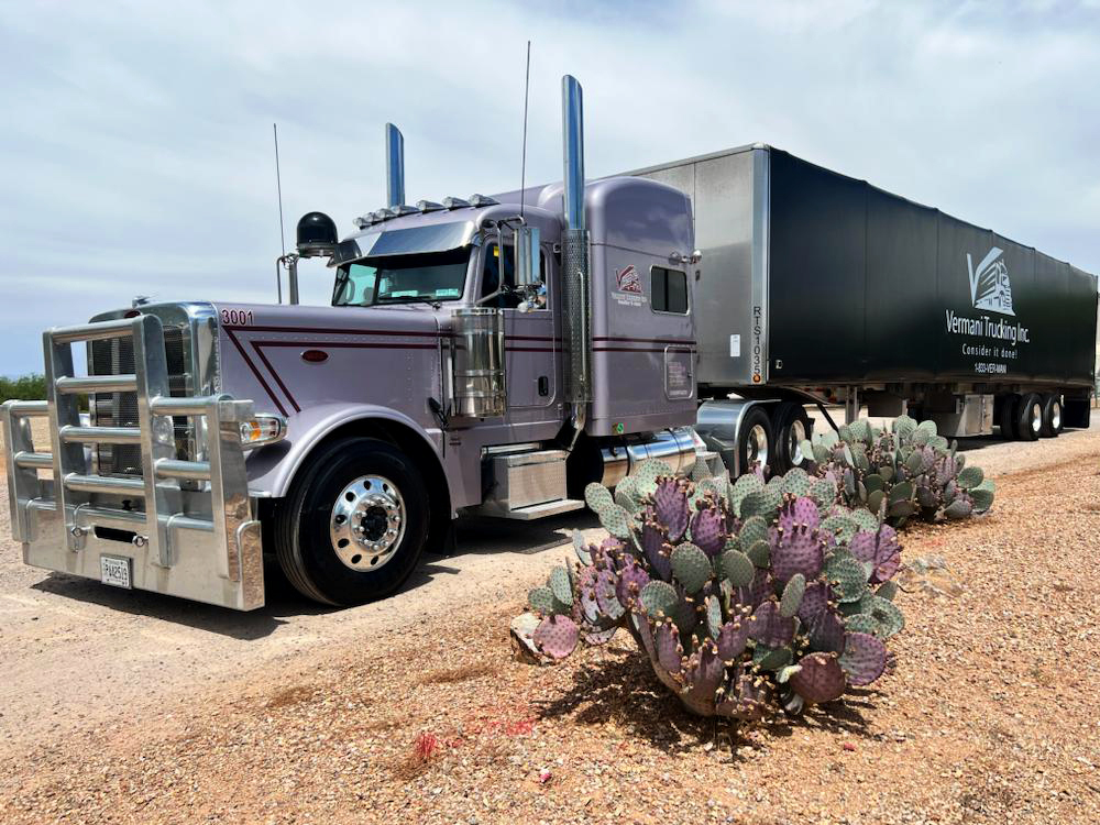 Silver Peterbilt semi-truck with black Varnani Trucking trailer parked on gravel with prickly pear cacti in foreground.