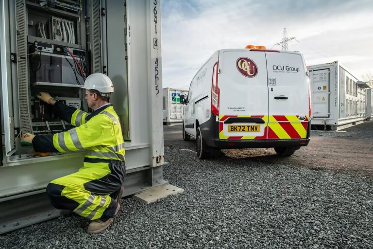 Technician in safety gear working on electrical equipment with OCU Group service van parked nearby at construction site.