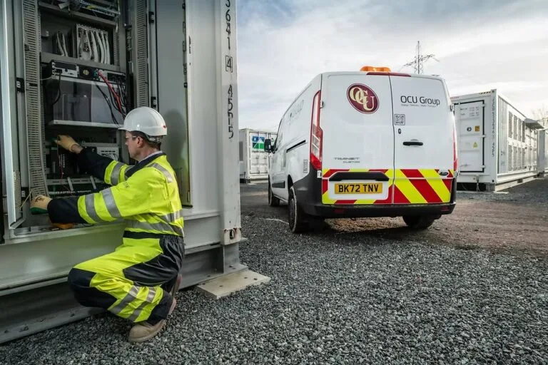 Technician in safety gear working on electrical equipment with OCU Group service van parked nearby at construction site.