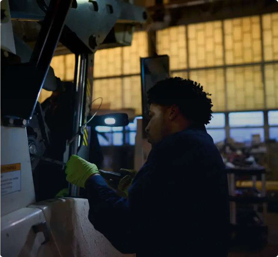 Worker in blue uniform with neon green gloves operating industrial machinery in a dimly lit warehouse.