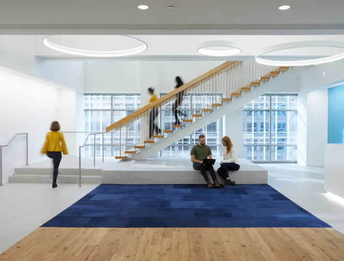 The lobby of an office building with people walking up and down the stairs and two people sitting while looking at a laptop.