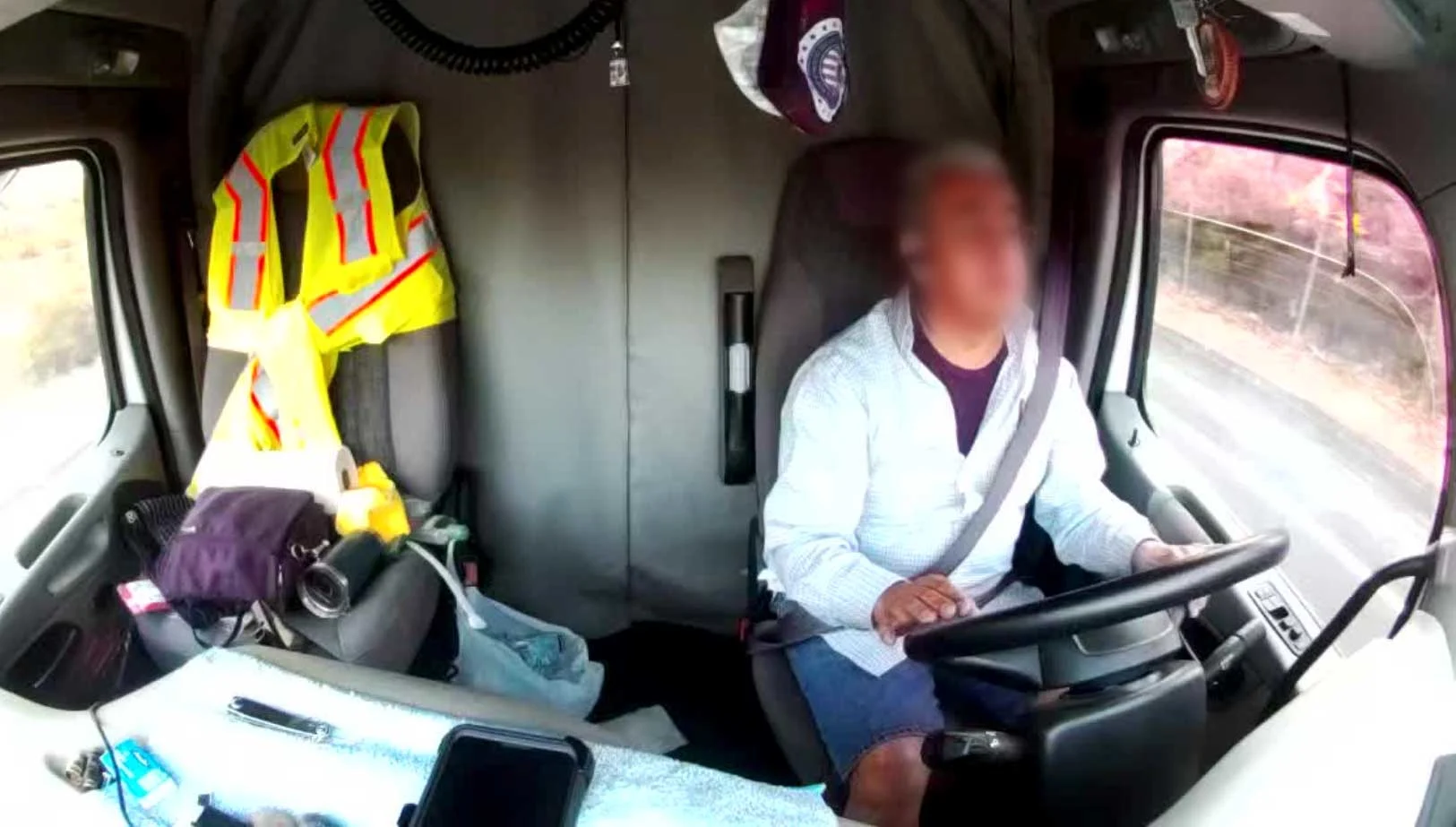 A driver at the wheel of a truck, passenger seat holds his belongings. Face is blurred. 