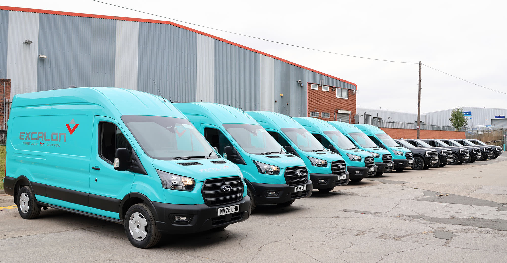 A fleet of teal Excalon-branded Ford Transit vans lined up outside an industrial building, with black vehicles behind them.