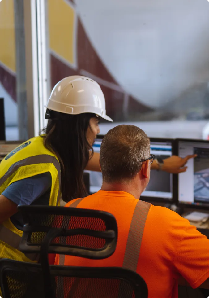 Construction professionals in safety gear reviewing plans on computer monitors at a worksite office.