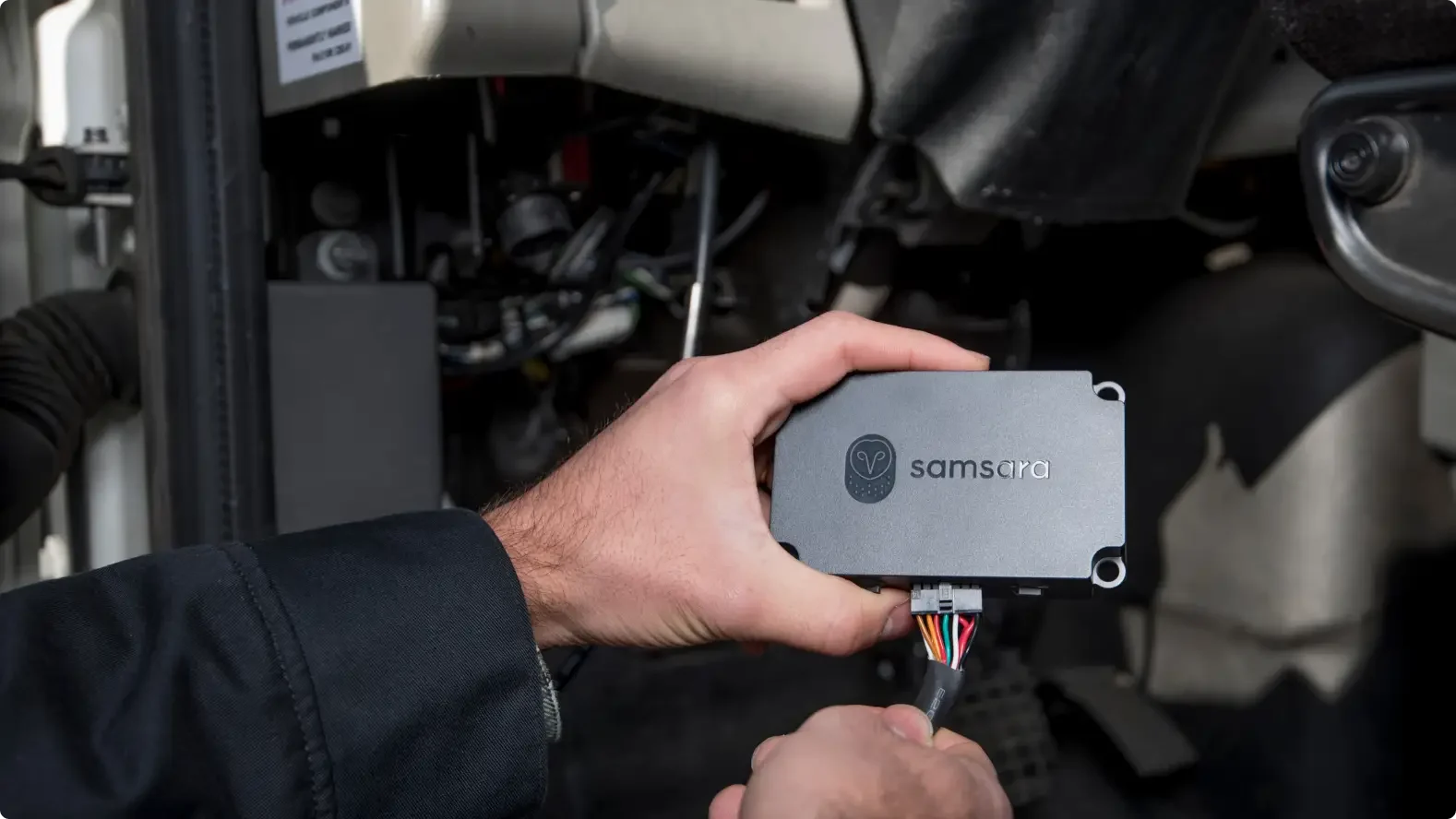 Hand installing a gray Samsara device with colored wires into a vehicle's dashboard area.