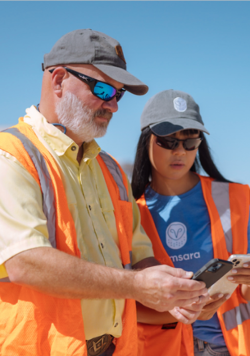 Two workers in orange safety vests and caps looking at mobile devices against a clear blue sky.