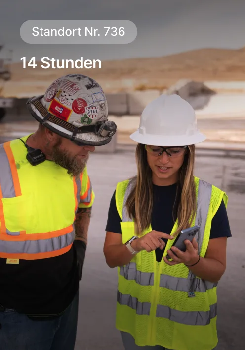 Zwei Bauarbeiter in Sicherheitswesten und Schutzhelmen überprüfen Notizen während Schicht in einer Wüstenumgebung.