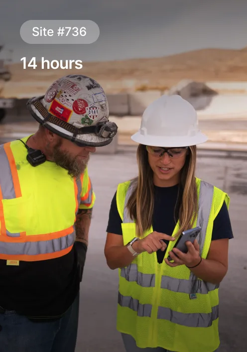 Two construction workers in safety vests and hard hats review notes at Site #736 during a 14-hour shift in a desert setting.