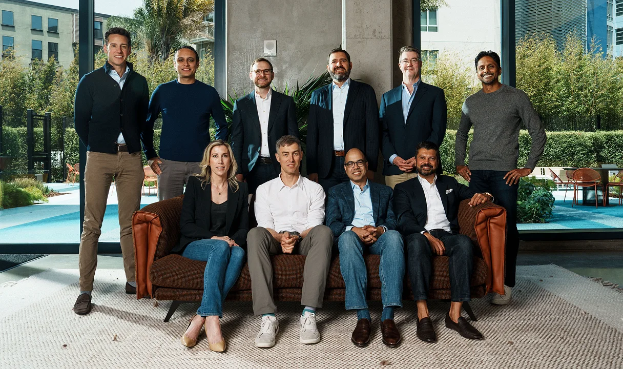 Group of professionals posed in modern office space with large windows, some seated on brown leather couch, others standing.