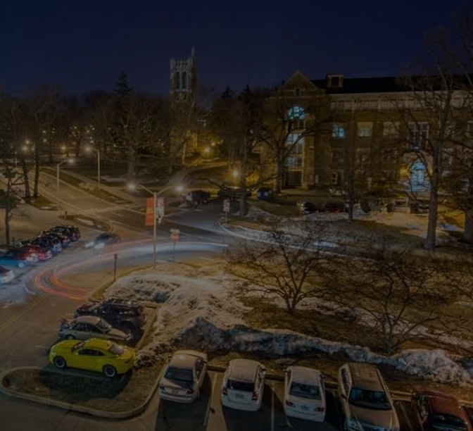 Night view of a college campus with brick buildings, snow-covered grounds, parked cars including a yellow vehicle