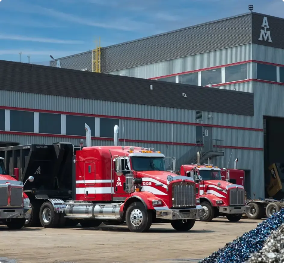 Camions rouges portant le logo AM garés devant un bâtiment industriel gris avec des bordures rouges sous un ciel bleu.