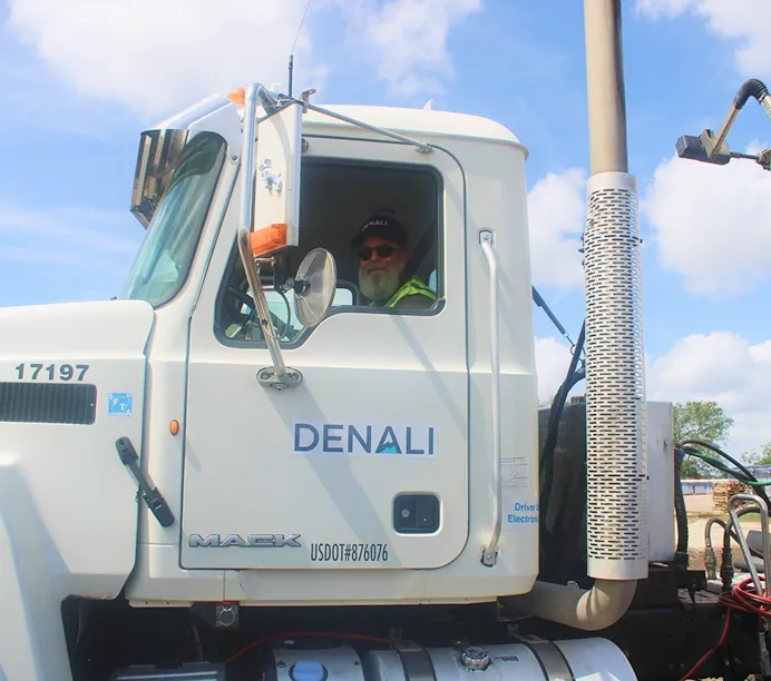 Driver in a white Mack truck cab labeled "DENALI" with chrome exhaust stack against blue sky.