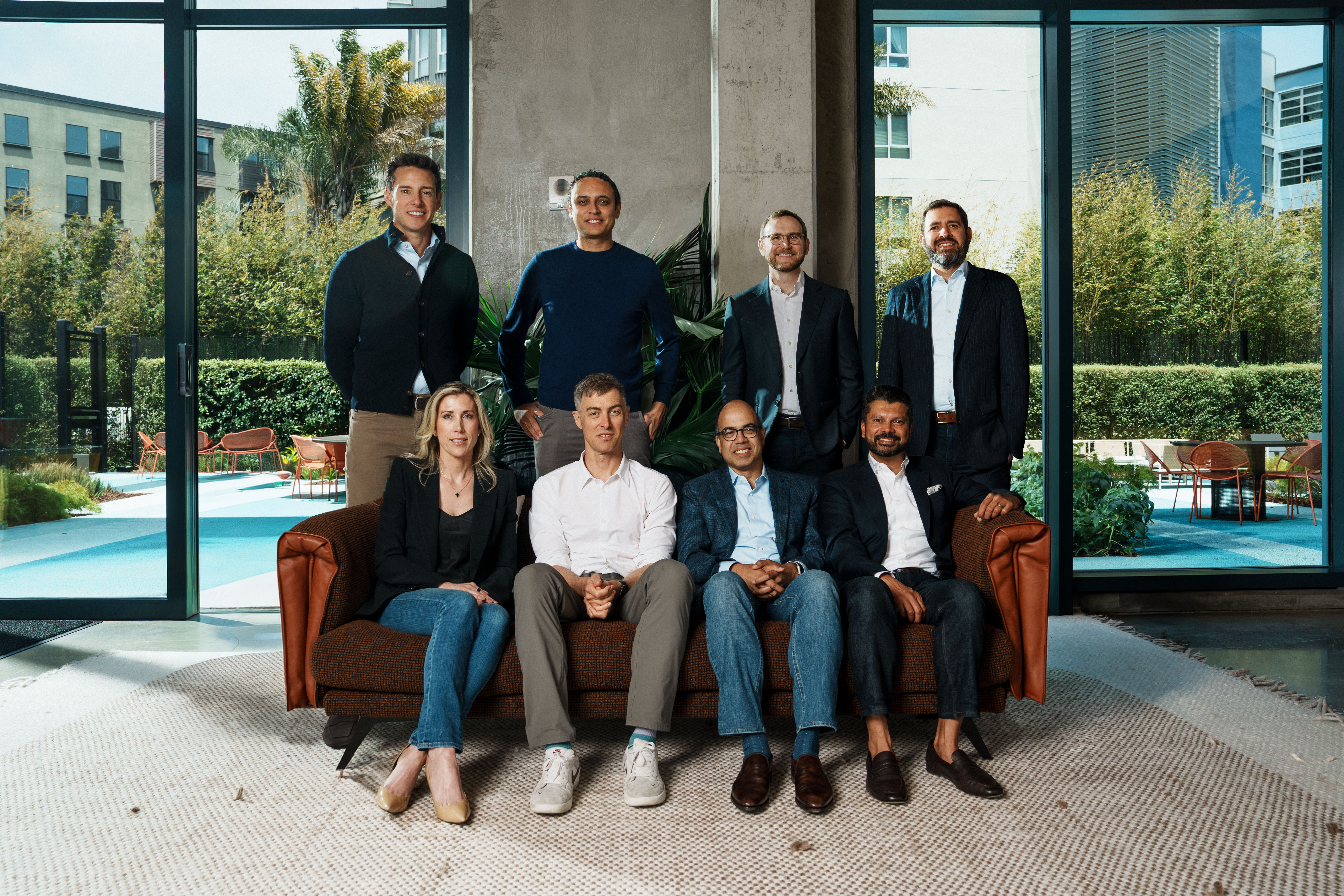 Eight professionals pose for a group photo indoors, with four seated on a brown sofa and four standing behind them.