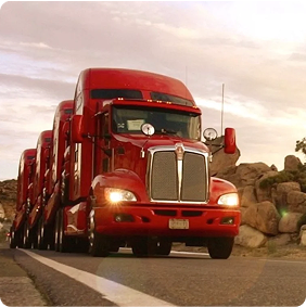 Fila de camiones rojos de carga pesada transitando por una carretera desértica con formaciones rocosas al atardecer.