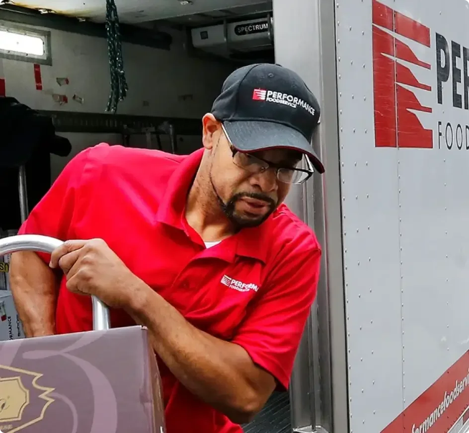 Delivery worker in red Performance shirt and cap loading items from a food service truck with PER branding.