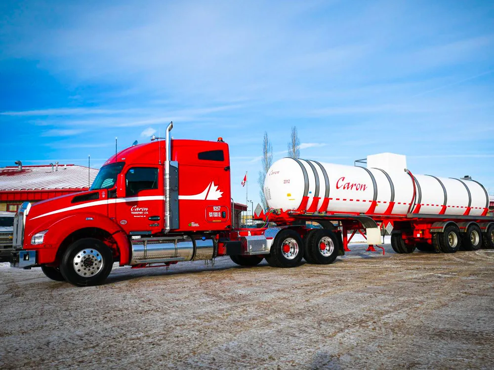 Camion-citerne Caron rouge vif avec une remorque cylindrique blanche garée sur un terrain de gravier sous un ciel bleu clair.