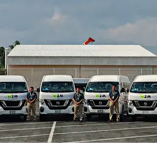 Fila de camionetas blancas con logo W2M y personal uniformado frente a un almacén con techo metálico bajo cielo nublado.