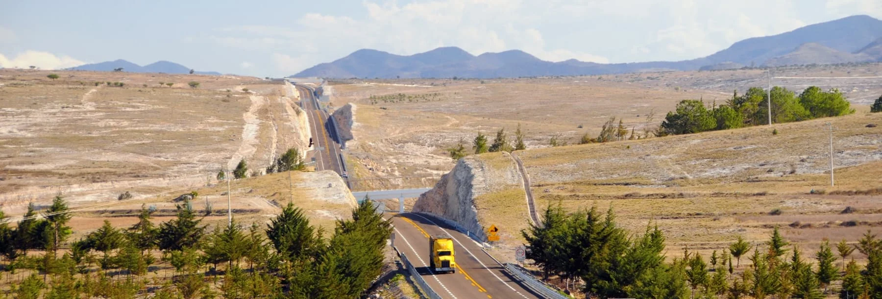 Yellow trucks drives along freeway next to grassy hills 