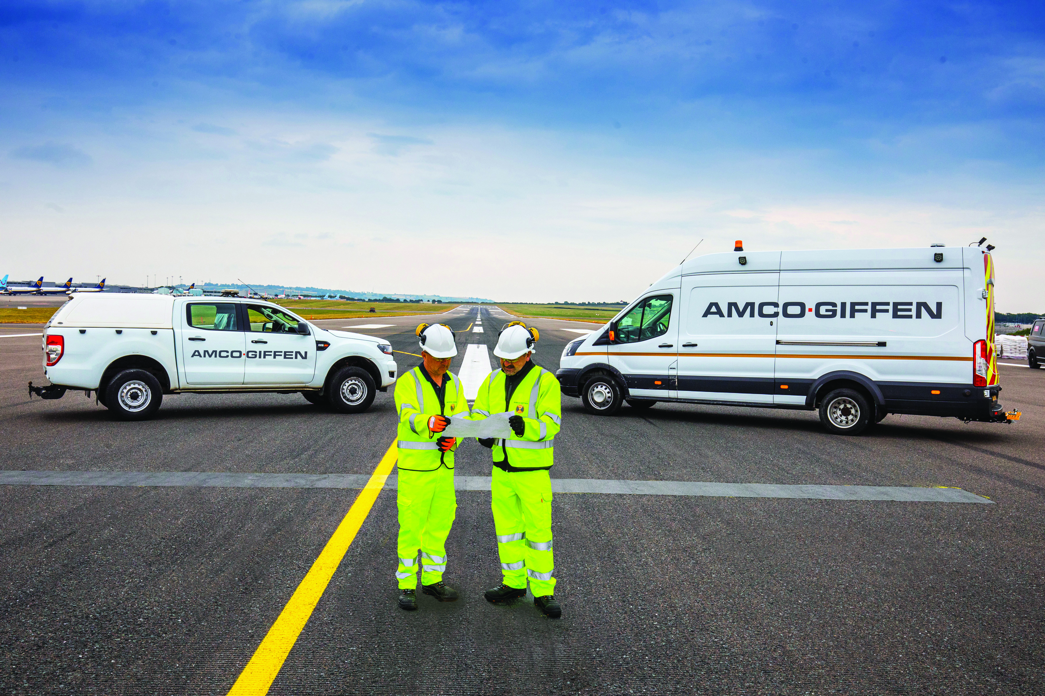 Two Amco Giffen workers in hi-vis gear review documents on an airport runway, flanked by branded white vehicles.