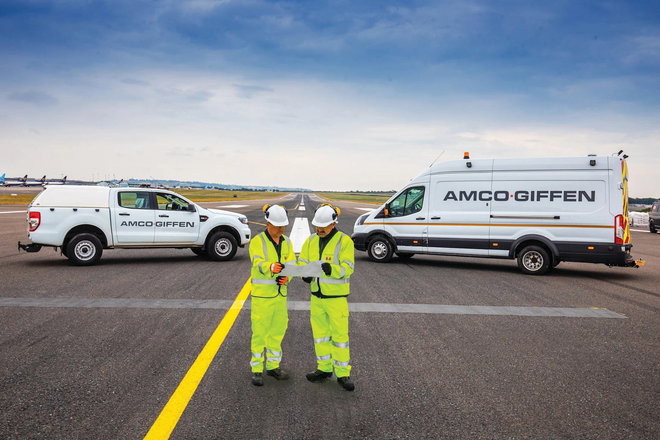 Two Amco Giffen workers in hi-vis gear review documents on an airport runway, flanked by branded white vehicles.