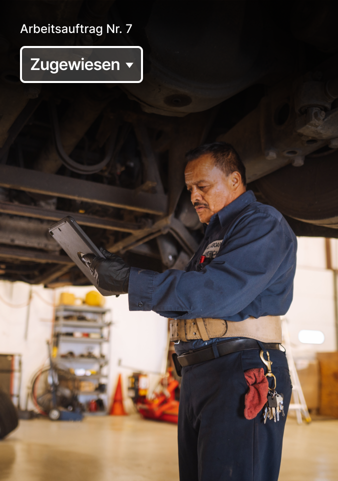 Mechanic in blue uniform examining vehicle undercarriage with diagnostic device, work order interface visible above.