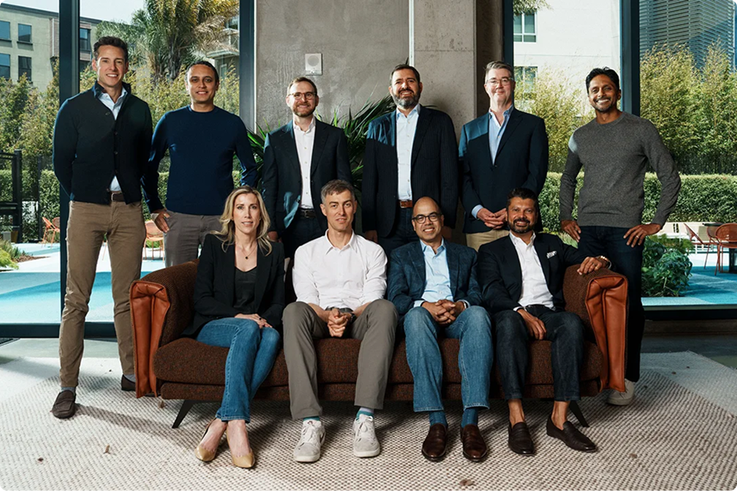 Group of professionals in business attire posed in modern office space with large windows overlooking greenery and pool.