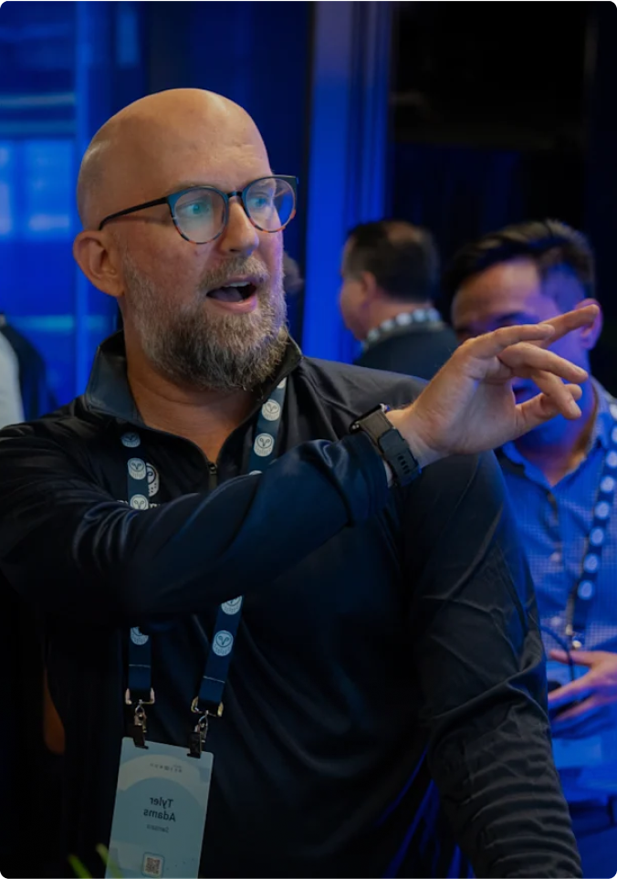 Bald man with beard and glasses gesturing while speaking at event, wearing lanyard and black shirt in blue-lit environment.