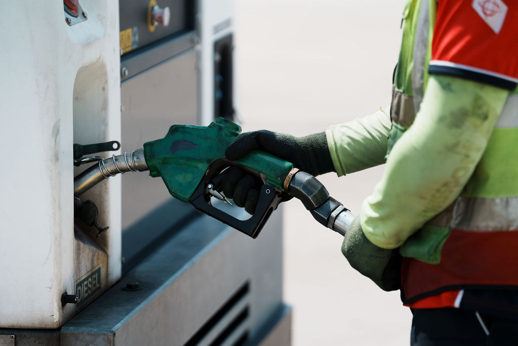 A worker in a red vest and green gloves holds a green diesel fuel nozzle at a pump.