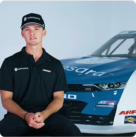 Piloto con uniforme negro y gorra junto a un auto de carreras azul con patrocinadores en un estudio fotográfico.