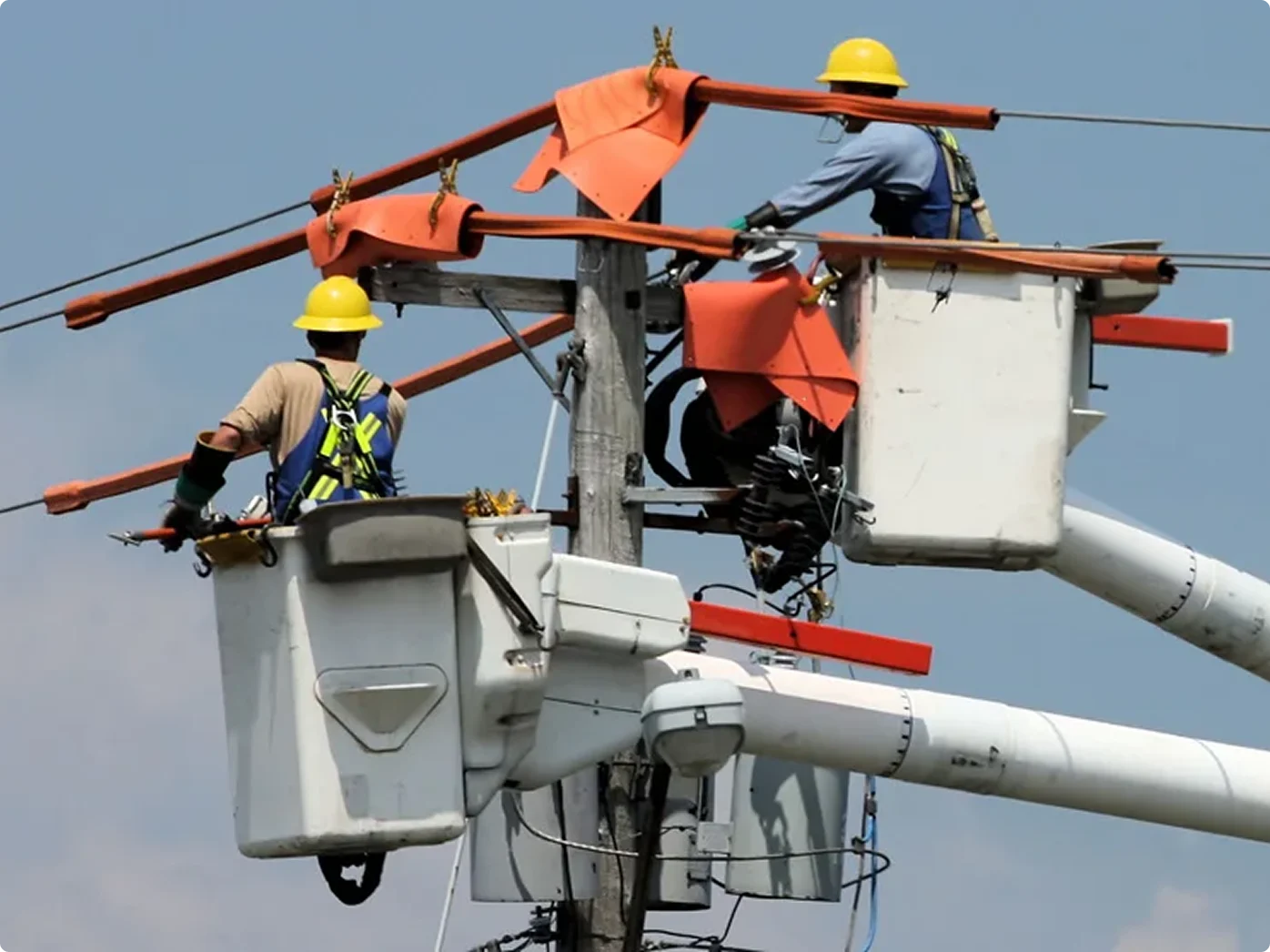 Utility workers in yellow hard hats repairing power lines from bucket trucks, with orange safety covers on equipment.