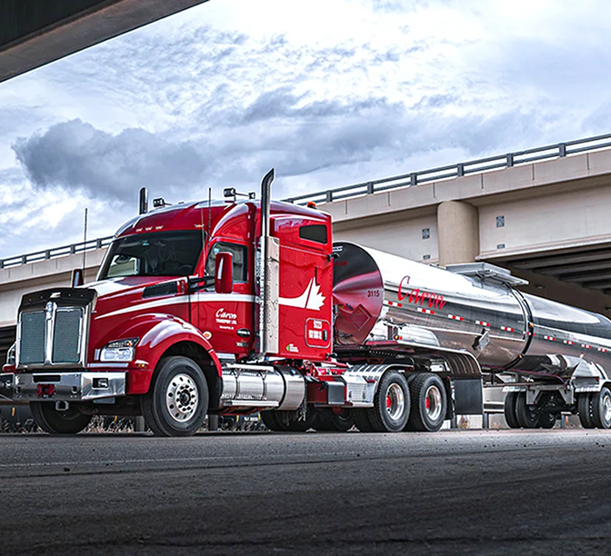 Red semi-truck with chrome tanker trailer parked under a highway overpass against cloudy blue sky.