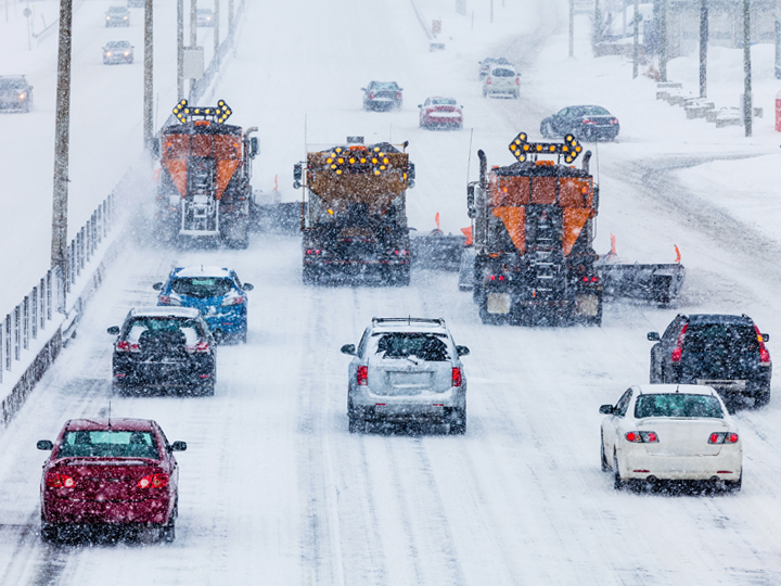 Snow plows clearing a highway during heavy snowfall with cars carefully navigating the snowy road conditions.