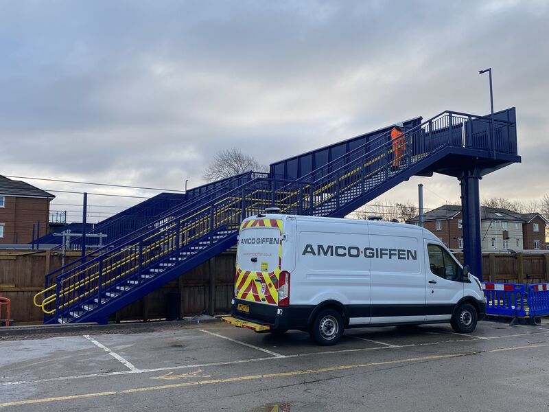 An Amco Giffen van parked beside a blue steel footbridge with yellow handrails at a railway station construction site.
