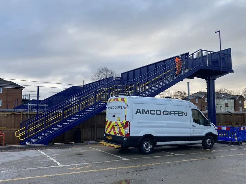 An Amco Giffen van parked beside a blue steel footbridge with yellow handrails at a railway station construction site.