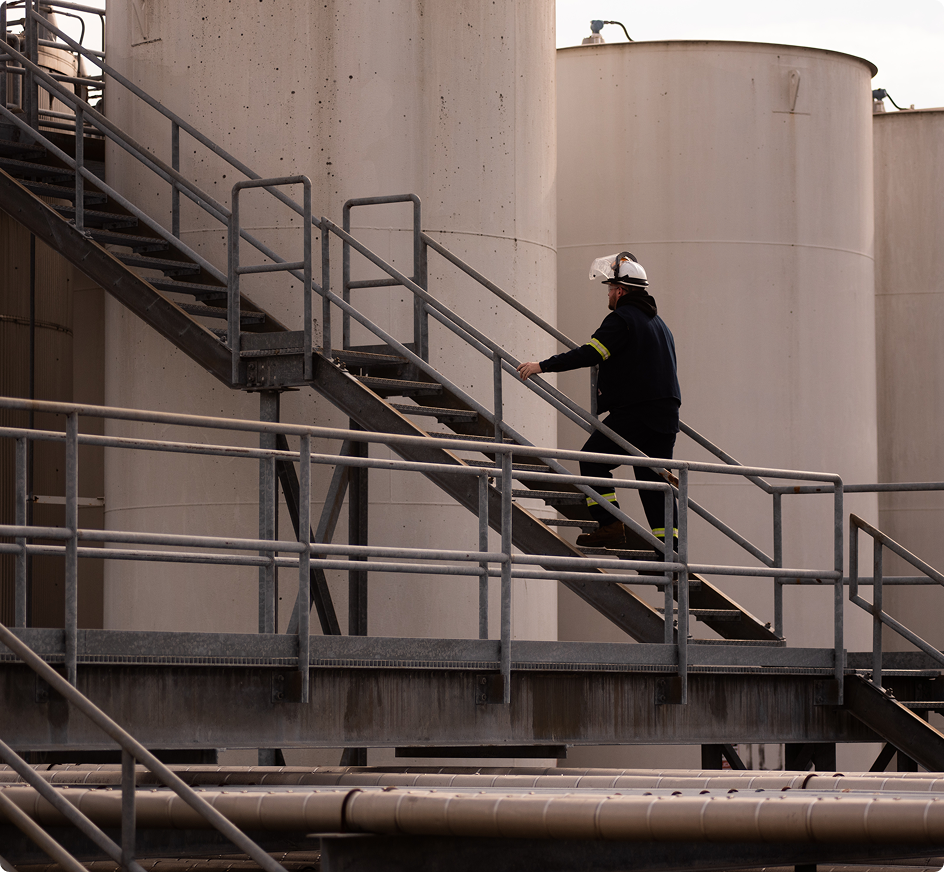 Worker in safety helmet climbing metal stairs at industrial facility with large beige storage tanks in background.